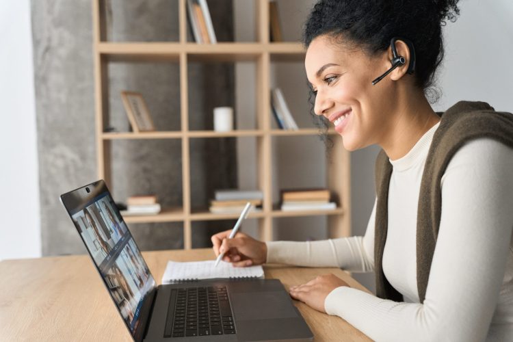 African American mixed race adult student wearing headset having virtual meeting online call educational webinar chatting at home office writing notes. Video e learning conference call on pc.
